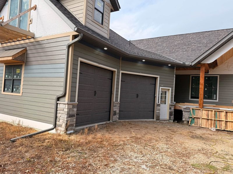 Grey house with dark brown garage doors, dark gutters and a downspout with an extension